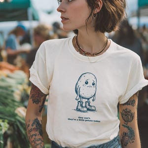 May include: Cream-colored t-shirt featuring a sad cartoon potato wearing boots. The text below the potato reads "they can't, they're a little potato today." The shirt is worn by a person at a farmer's market.