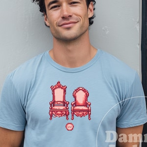 May include: Light blue t-shirt with a pixelated design of two ornate pink chairs. The shirt has the text "Chambray" at the top and "Dammit Judy" at the bottom. The person is smiling.