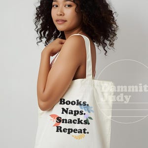 以下が含まれることがあります： オフホワイトのキャンバス地のトートバッグ。黒字で「Books, Naps, Snacks, Repeat」と書かれ、花の装飾が施されています。バッグには2本のショルダーストラップがあり、人が持っています。