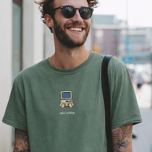 Può includere: T-shirt verde oliva con una piccola grafica di computer e le parole "vibe coding" stampate sotto. La persona indossa occhiali da sole e una catena d'oro. La persona sorride e ha tatuaggi sul braccio.