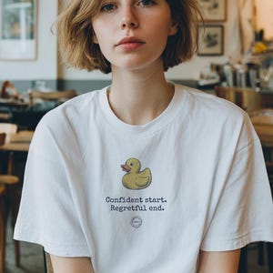 May include: White t-shirt with a yellow cartoon duck and the text "Confident start. Regretful end." The shirt is worn by a person with short brown hair, in a cafe setting.