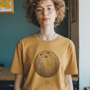 May include: Mustard-coloured t-shirt with a detailed illustration of a bowling ball. The graphic is centered on the shirt. The person is wearing glasses and a silver necklace. The background includes a teal wall and a framed picture.