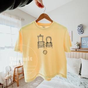 May include: A yellow t-shirt with a black and white illustration of two ornate chairs. The shirt has a crew neck and short sleeves. A small circular graphic is below the chair illustration.