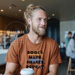 May include: A brown t-shirt with the words "BOOKS, NAPS, SNACKS, REPEAT" in a stacked design. The person in the image is holding a coffee cup with a white lid. The image is taken indoors.