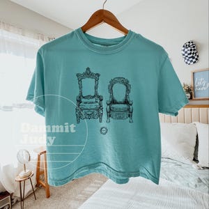 May include: A teal short-sleeved t-shirt featuring a black graphic of two ornate chairs. The shirt is hanging on a wooden hanger. The text "Dammit Judy" is visible on the shirt.