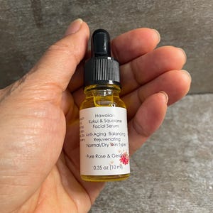 May include: A small glass bottle of Hawaiian Kukui & Squalane Facial Serum. The bottle has a black dropper top and a white label with text and a floral design. The label indicates the serum is for normal/dry skin types and contains Pure Rose & Geranium. The bottle contains 10 ml.