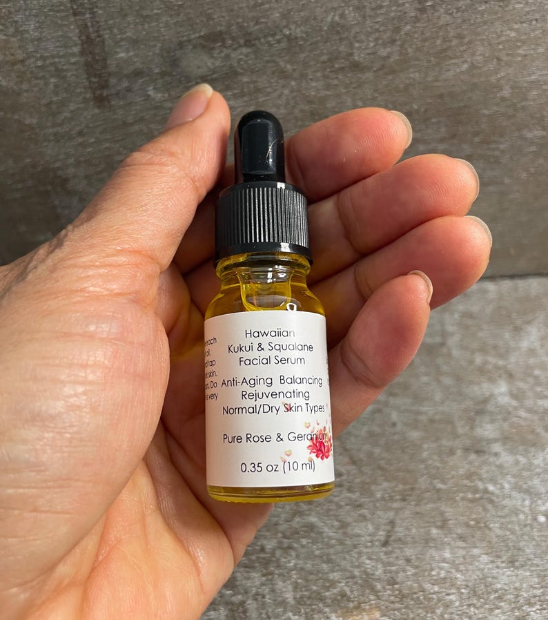 May include: A small glass bottle of Hawaiian Kukui & Squalane Facial Serum. The bottle has a black dropper top and a white label with text and a floral design. The label indicates the serum is for normal/dry skin types and contains Pure Rose & Geranium. The bottle contains 10 ml.