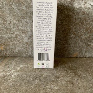 May include: A white product box detailing a facial serum made with Hawaiian Kukui Nut and Olive Squalane Oils. The text highlights anti-ageing, moisturising, and protective qualities for all skin types.