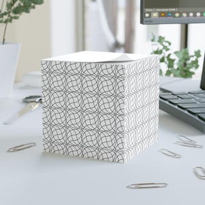 May include: A white cube notepad featuring a black wavy line design. The notepad sits on a white desk, surrounded by silver paper clips, scissors, and a laptop.