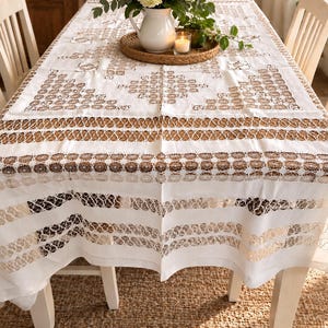 May include: A white rectangular tablecloth featuring intricate brown lace inserts and geometric cutwork patterns. The fabric displays horizontal bands of circular lace motifs and solid white sections, creating a textured appearance across the surface.