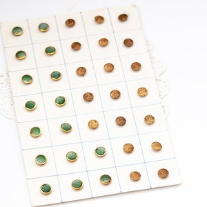 May include: A collection of vintage buttons arranged on a grid. Some buttons are green with gold rims, while others are textured and bronze-colored. The buttons are displayed on a white card with a grid pattern, and a white lace doily is visible.