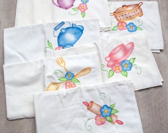 May include: A collection of white kitchen towels, each adorned with hand-painted watercolor designs. The designs include teapots, a saucepan, a tea cup, and utensils, all surrounded by colorful flowers. The towels are arranged on a light gray surface.