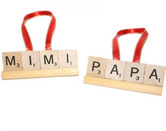 May include: Two wooden ornaments with red ribbon hangers. One spells "MIMI" and the other "PAPA" using Scrabble tiles. The text "Custom 1-4 Letters" is in green script below the ornaments. The background is white.