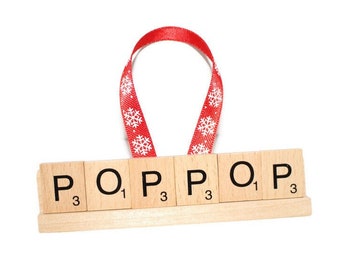 May include: Wooden ornament spelling "POP" with Scrabble tiles, featuring a red ribbon with white snowflake patterns. The letters and numbers are printed in black. The ornament is on a white background.