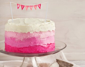 May include: A layered cake with a gradient of pink frosting, topped with white frosting and a banner that reads "Olivia." The cake sits on a glass cake stand. A cake server rests on a white cloth.