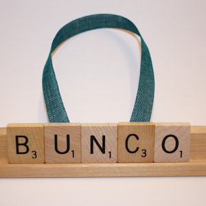 May include: Wooden tile sign spelling "BUNCO" with a teal ribbon handle. The tiles are arranged on a wooden base. The letters and numbers are black. The background is white.