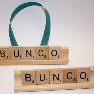 May include: Two wooden Bunco game tile holders. One has a teal ribbon handle. Each holder displays the word "BUNCO" spelled out with individual letter tiles. The tiles are light brown with black letters and numbers.