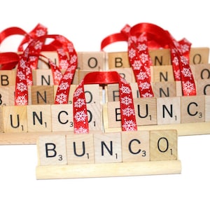 May include: Wooden Bunco game pieces with red snowflake ribbon. The game pieces are arranged in a tiered display, spelling out "Bunco" with individual letter tiles. The ribbon adds a festive touch.