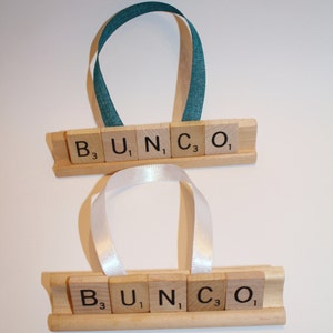 May include: Two wooden ornaments with the word "BUNCO" spelled out in Scrabble tiles. Each ornament has a ribbon for hanging, one teal and one white. The letters are on a wooden base.