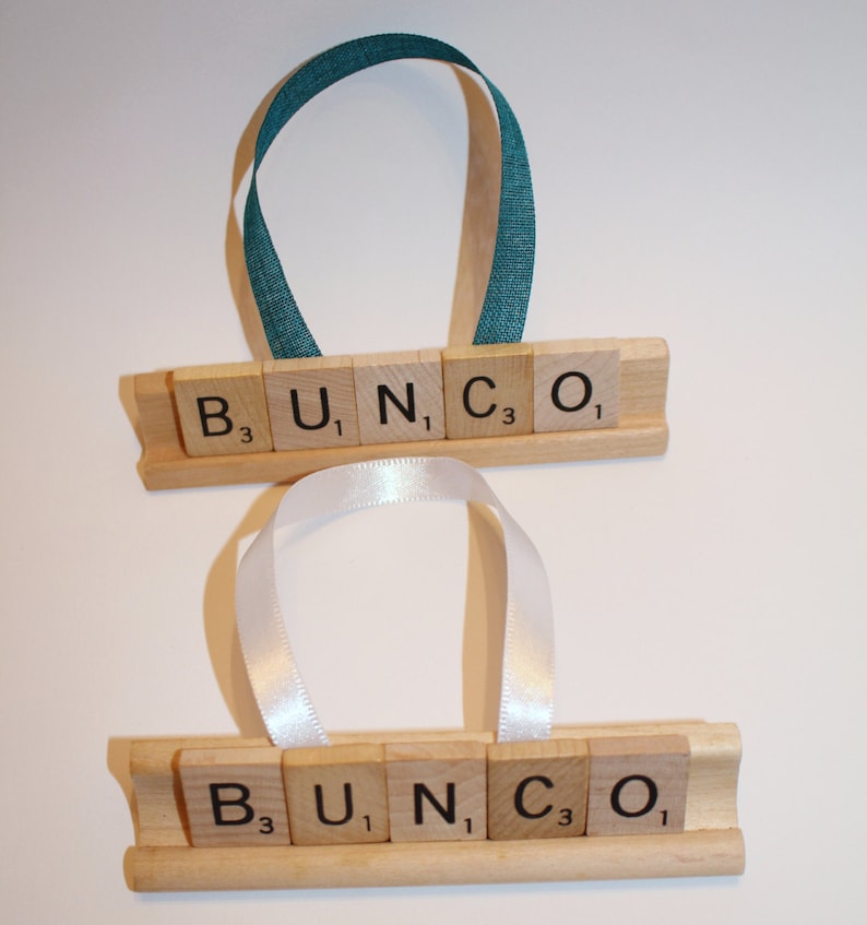 May include: Two wooden ornaments with the word "BUNCO" spelled out in Scrabble tiles. Each ornament has a ribbon for hanging, one teal and one white. The letters are on a wooden base.