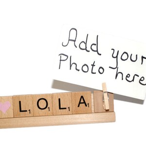 May include: A wooden photo holder with the word "LOLA" spelled out in Scrabble tiles. A pink heart tile is on the left. A white card with the text "Add your photo here" is attached to the holder with a wooden clothespin.