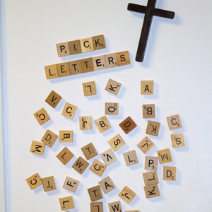 May include: A white surface with a wooden cross and a collection of letter tiles spelling out "PICK LETTERS". The tiles are scattered around the cross.