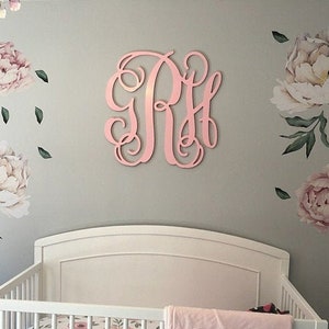 May include: A white crib with a pink floral crib sheet and a pink blanket. The wall behind the crib is decorated with pink floral decals and a large pink wooden monogram.