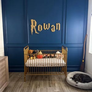 May include: A gold-colored crib filled with stuffed animals sits in front of a dark blue wall. The name "Rowan" is displayed in gold lettering above the crib. A black dog rests in a gray dog bed on the floor.