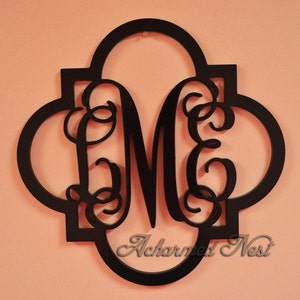 May include: Black wooden monogram with the letters "ME" in a decorative script font. The monogram is in the shape of a quatrefoil and has a cutout design. The text "Acharmed Nest" is visible at the bottom of the monogram.