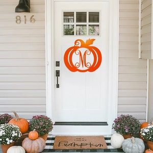 May include: A white front door with a pumpkin-shaped monogram on it. The door has a black door handle and a black light fixture above it. There is a welcome mat with the text "Hey There Pumpkin" on it. There are pumpkins and mums on the porch.