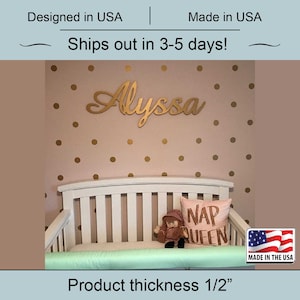May include: Gold wooden name sign with the name "Alyssa" in cursive script. The sign is mounted on a pink wall with gold polka dots. A white crib with a green bumper and a pillow with the text "NAP QUEEN" is in the foreground. The sign is 1/2 inch thick.
