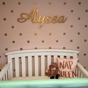 May include: A white crib with a green bumper pad and a pink pillow with the text "NAP QUEEN". A doll with brown hair and a pink dress is sitting on the crib. The wall behind the crib is pink with gold polka dots and a gold cursive name sign that says "Alyssa".