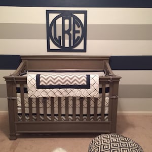 May include: A brown wooden crib with a white and gray chevron patterned crib bedding. The crib is in front of a wall with alternating navy blue and gray horizontal stripes. A wooden sign with the letters "GRE" in a circle is hanging on the wall.