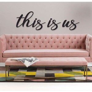 May include: A pink tufted sofa and matching ottoman with a black wooden sign that reads "this is us" hanging on the wall above the sofa.