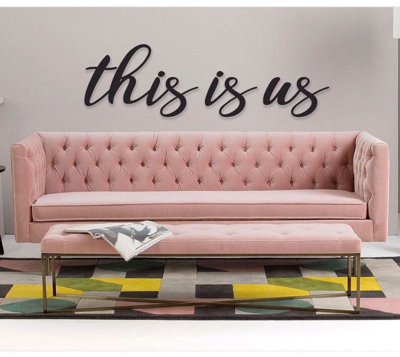 May include: A pink tufted sofa and matching ottoman with a black wooden sign that reads "this is us" hanging on the wall above the sofa.
