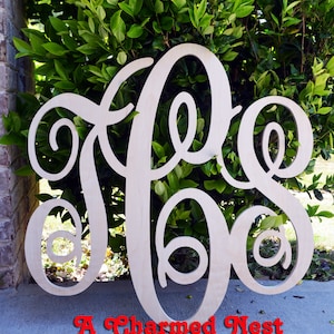 May include: A large, unfinished wooden monogram in the shape of the letters "J.E.O."  The monogram is standing upright against a backdrop of green foliage. The text "A Charmed Nest" is visible at the bottom of the image.