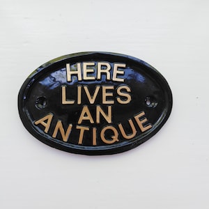 May include: Black oval sign with gold lettering that reads "Here Lives An Antique".