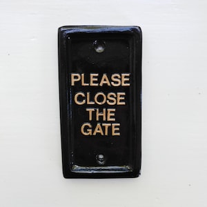 May include: Black metal sign with gold lettering that reads "PLEASE CLOSE THE GATE".