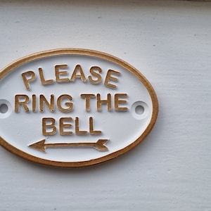 Könnte beinhalten: Ein weißes ovales Schild mit goldener Umrandung und einem Pfeil, der nach rechts zeigt. Auf dem Schild steht "Please Ring The Bell".