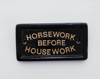 Może przedstawiać: Czarny metalowy znak z pozłacanymi literami, na którym widnieje napis "Horsework Before Housework".