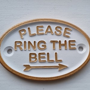 Könnte beinhalten: Ein weißes ovales Schild mit goldener Umrandung und dem Text "PLEASE RING THE BELL" mit einem goldenen Pfeil, der nach rechts zeigt.