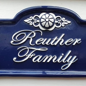 Puede incluir: Una placa de n&uacute;mero de casa de cer&aacute;mica azul y blanca con el texto "Reuther Family" en letras cursivas blancas.