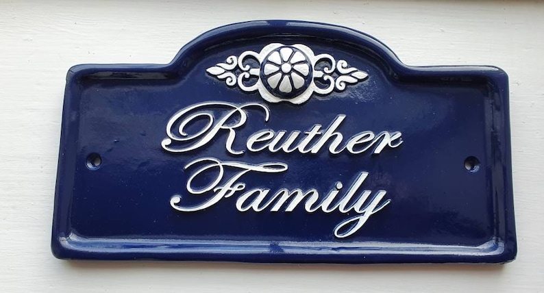 Puede incluir: Una placa de n&uacute;mero de casa de cer&aacute;mica azul y blanca con el texto "Reuther Family" en letras cursivas blancas.
