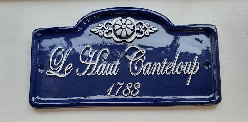 Puede incluir: Un letrero de esmalte azul y blanco con el texto "Le Haut Canteloupe 1783". El letrero tiene un dise&ntilde;o floral decorativo en la parte superior.