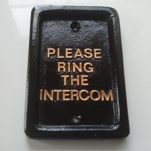 Op de afbeelding: Zwart gietijzeren bord met gouden letters met de tekst "PLEASE RING THE INTERCOM".