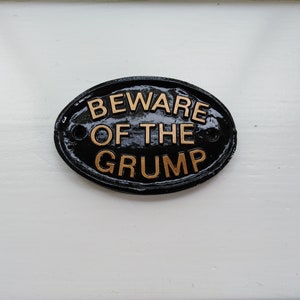 Może przedstawiać: Czarny owalny znak z napisem "Beware of the Grump" w kolorze złotym.