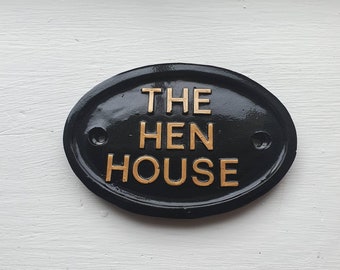 Op de afbeelding: Zwart ovaal bord met gouden letters met de tekst "THE HEN HOUSE".