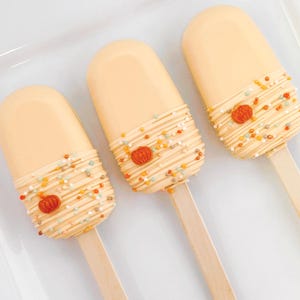 May include: Three cake pops shaped like ice cream bars on wooden sticks. Each treat is coated in a smooth, light orange icing with a drizzle of matching icing, small multi-colored sprinkles, and a single miniature orange pumpkin decoration.