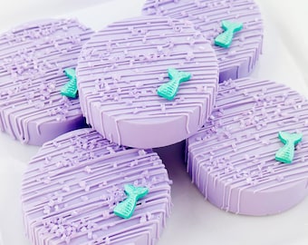 May include: Several round, lavender-colored treats with diagonal white lines and sprinkles. Each treat is decorated with a small, turquoise mermaid tail. The treats are arranged on a white plate, creating a whimsical and thematic dessert.