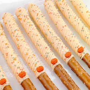 May include: A row of pretzel rods dipped in light orange icing, drizzled with additional orange icing, and decorated with small, multi-colored sprinkles and a single orange pumpkin-shaped candy at the base of the coated section.